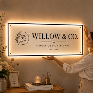 K&ouml;nnte beinhalten: Ein rechteckiges Wand-Schild mit schwarzem Rahmen und Hintergrundbeleuchtung. Die wei&szlig;e Oberfl&auml;che zeigt ein florales Strichm&auml;nnchen-Symbol, den Text "WILLOW & CO.", "FLORAL DESIGN & CAF&Eacute;" und "EST. 2025" mit einem kleinen Herz unter dem Namen.