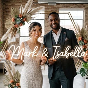 May include: A smiling couple holds a neon sign reading "Mark & Isabella." The sign emits a warm light. The background includes floral arrangements and a geometric backdrop, possibly for a wedding or event.