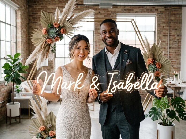May include: A smiling couple holds a neon sign reading "Mark & Isabella." The sign emits a warm light. The background includes floral arrangements and a geometric backdrop, possibly for a wedding or event.