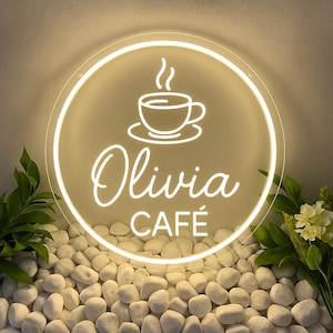 May include: A circular neon sign for "Olivia Cafe", featuring a coffee cup illustration. The sign glows with a white light, displaying the cafe's name in cursive script and the word "CAFE" below. The sign is set against a backdrop of white pebbles and foliage.