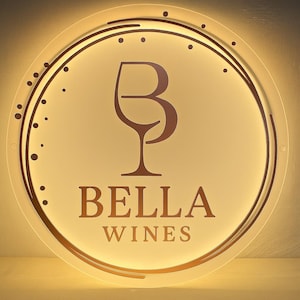 K&ouml;nnte beinhalten: Ein kreisf&ouml;rmiges, hinterleuchtetes Wandschild mit dem Text BELLA WINES in einer Serifenschrift. &Uuml;ber dem Text befindet sich eine stilisierte Grafik, die die Buchstaben B und P zu einem Weinglas kombiniert, erg&auml;nzt durch geschwungene Linien.