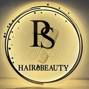 K&ouml;nnte beinhalten: Ein kreisf&ouml;rmiges, beleuchtetes Schild mit den schwarzen Buchstaben PS in einer Serifenschrift, hinter denen sich die schwachen Umrisse einer Schere und eines Kamms befinden. Der Text HAIR&BEAUTY steht in Schwarz unter den Buchstaben.