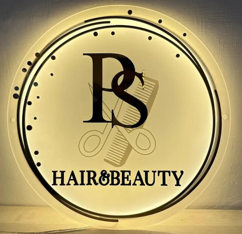 K&ouml;nnte beinhalten: Ein kreisf&ouml;rmiges, beleuchtetes Schild mit den schwarzen Buchstaben PS in einer Serifenschrift, hinter denen sich die schwachen Umrisse einer Schere und eines Kamms befinden. Der Text HAIR&BEAUTY steht in Schwarz unter den Buchstaben.