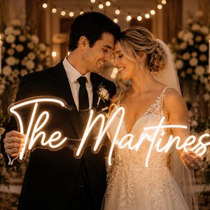 May include: A couple smiles, holding a neon sign that reads "The Martinez." The sign is in a cursive font and glows with a warm light. The bride wears a white wedding dress, and the groom is in a black suit. The background features candles and floral arrangements.