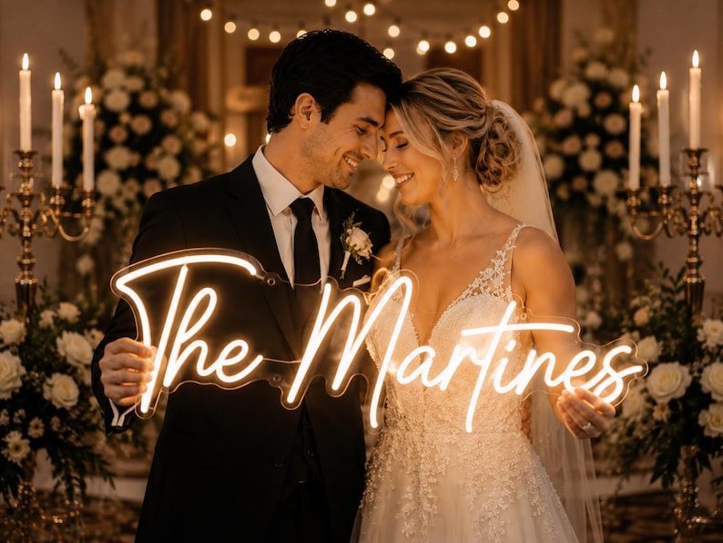 May include: A couple smiles, holding a neon sign that reads "The Martinez." The sign is in a cursive font and glows with a warm light. The bride wears a white wedding dress, and the groom is in a black suit. The background features candles and floral arrangements.