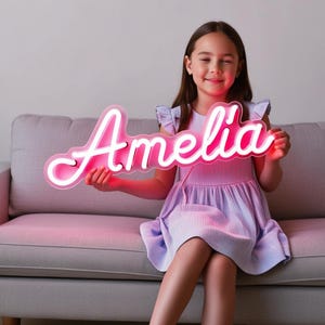 May include: A young person holding a pink neon sign with the name "Amelia" written in white. The sign is lit up and casting a pink glow.