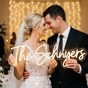 May include: A smiling couple holds a neon sign reading "The Schnyers" at a wedding. The bride wears a white gown and veil, the groom a black suit and tie. The background features floral arrangements and candlelit decorations. The sign is in a cursive font.