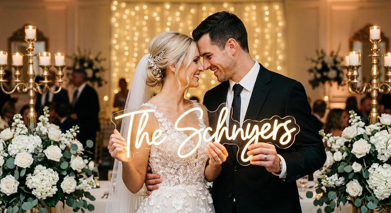 May include: A smiling couple holds a neon sign reading "The Schnyers" at a wedding. The bride wears a white gown and veil, the groom a black suit and tie. The background features floral arrangements and candlelit decorations. The sign is in a cursive font.