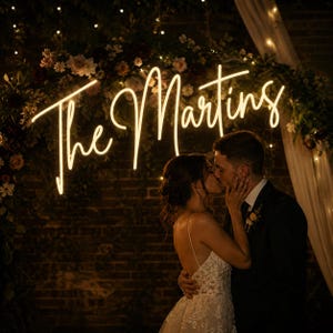 May include: A neon sign that reads "The Martins" in a cursive font, illuminated against a brick wall backdrop. A couple is kissing in front of the sign, with floral arrangements and string lights adding to the romantic ambiance.