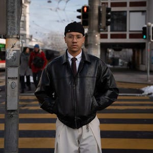 Op de afbeelding: Een persoon met een zwarte leren jas, een wit overhemd en een crèmekleurige broek staat op een zebrapad. De persoon draagt ook een zwarte muts, een bril en een bordeauxrode stropdas. Op de achtergrond zijn gebouwen en een verkeerslicht te zien.