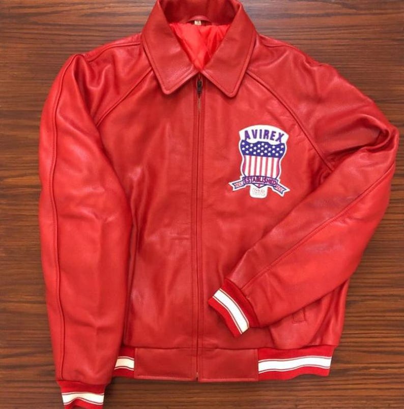Front view of bright red Avirex Icon leather bomber jacket featuring premium genuine leather construction, classic collared design, and iconic embroidered Avirex USA shield patch on the chest. This retro varsity bomber jacket blends vintage hip hop fashion, Y2K streetwear, and luxury urban style with a sleek zip-up silhouette and striped ribbed cuffs and hem. Perfect men’s leather bomber jacket for biker fashion, casual winter outfits, oversized street style, and trendy retro outerwear looks.