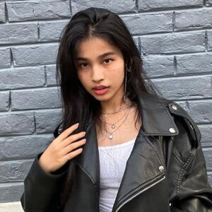 May include: A black leather jacket with silver zips and buttons. The jacket is worn over a white top and layered necklaces. The background is a grey brick wall.