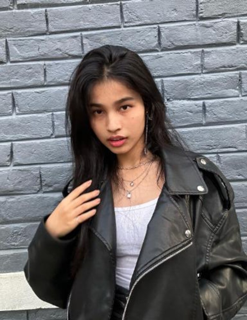 May include: A black leather jacket with silver zips and buttons. The jacket is worn over a white top and layered necklaces. The background is a grey brick wall.