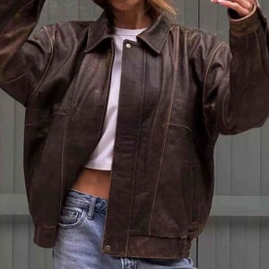 Könnte beinhalten: Braune Lederjacke mit klassischem Kragen und Knopfverschluss. Die Jacke hat eine leicht übergroße Passform und einen gerippten Saum und Bündchen. Die Jacke wird über einem weißen T-Shirt und einer blauen Jeans getragen.