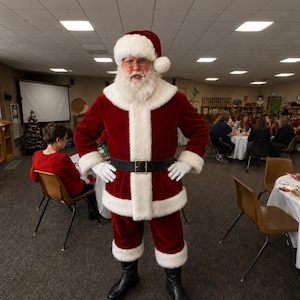 Puede incluir: Una persona vestida con un traje tradicional de Papá Noel de terciopelo rojo con ribetes de piel sintética blanca, gorro a juego, cinturón de cuero negro con hebilla dorada, guantes blancos y botas negras, con una larga barba blanca y gafas.