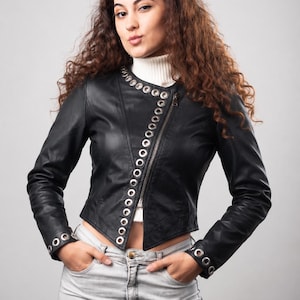 May include: A black leather jacket featuring a diagonal zip closure and silver-toned metal grommets lining the front opening and cuffs. The jacket is worn over a white ribbed turtleneck jumper paired with light grey denim jeans.
