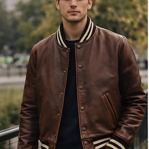 Peut inclure: Veste de bombardier en cuir marron avec col et poignets côtelés à rayures crème et marron. La veste a une fermeture à boutons et deux poches avant. Le modèle porte une chemise noire et un jean bleu.