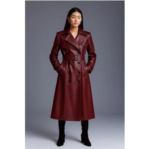 May include: A full-length burgundy trench coat with a double-breasted design, a belt, and a notched collar. The coat has a classic silhouette and is made of a smooth, leather-like material. The model is wearing black boots.