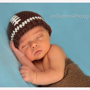 May include: A newborn baby wearing a brown and white crocheted football hat sleeps on a blue blanket. The baby is wearing a brown knitted wrap.