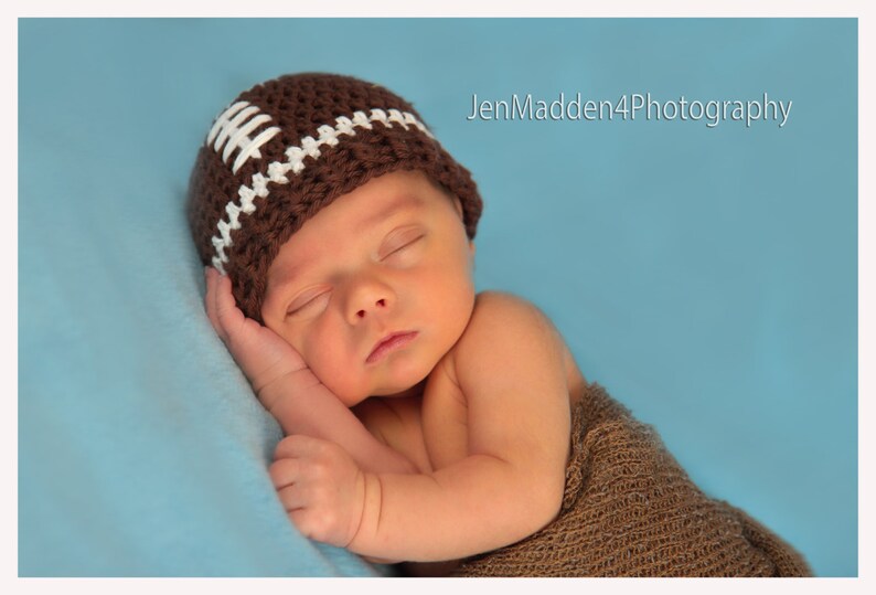 May include: A newborn baby wearing a brown and white crocheted football hat sleeps on a blue blanket. The baby is wearing a brown knitted wrap.