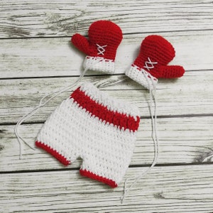 May include: A white crocheted baby romper with red trim and two red crocheted boxing gloves.