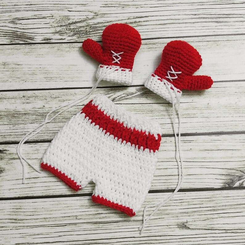 May include: A white crocheted baby romper with red trim and two red crocheted boxing gloves.