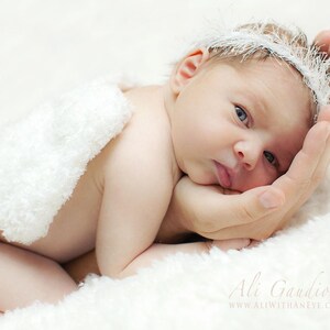 May include: A newborn baby wearing a white feathered headband and a white fluffy blanket, resting on a hand.