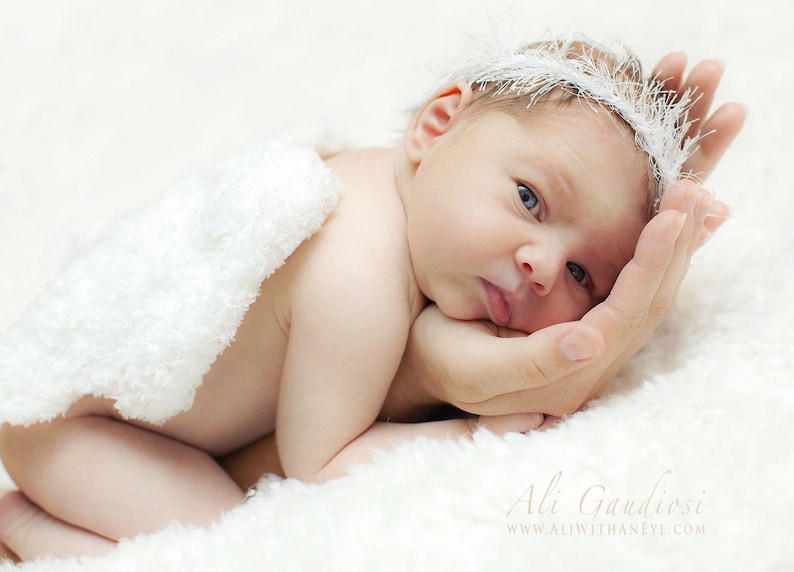 May include: A newborn baby wearing a white feathered headband and a white fluffy blanket, resting on a hand.