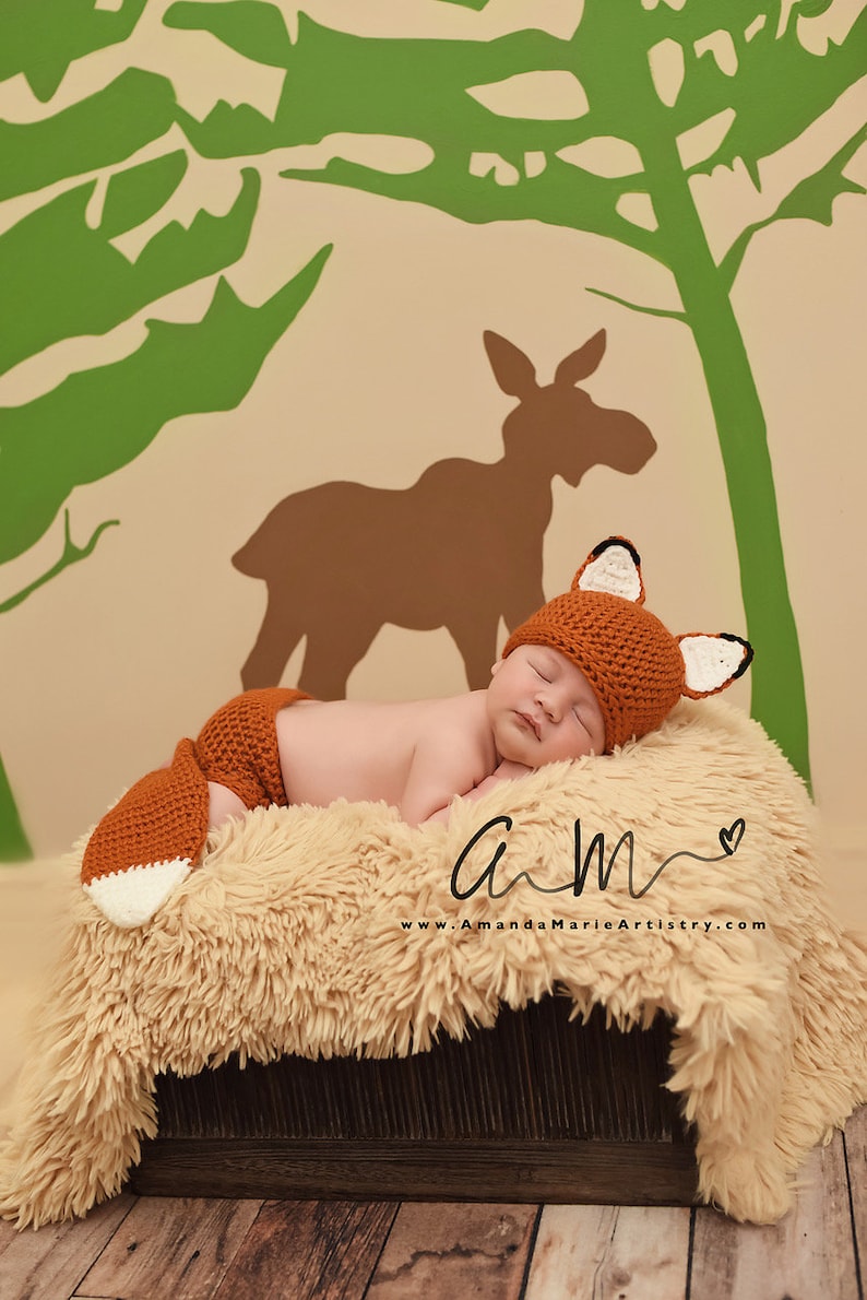 May include: A newborn baby wearing a crocheted fox hat and matching pants sleeps on a fluffy tan rug. The background features a green and brown silhouette of a moose and trees.