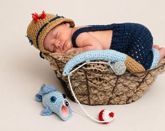 May include: A crocheted tan bucket hat with a red tassel, a dark blue overall set, and a light blue crocheted fishing rod with a matching fish toy. The set is displayed in a woven basket, featuring a small red and white crocheted bobber.
