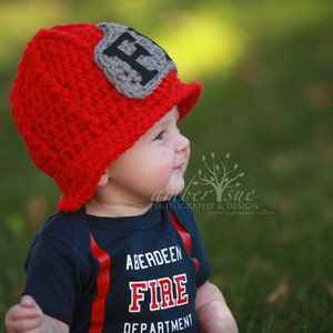 May include: A baby wearing a red crocheted firefighter hat with a gray patch that says "F" and a blue shirt that says "Aberdeen Fire Department".