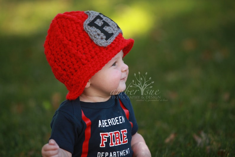 May include: A baby wearing a red crocheted firefighter hat with a gray patch that says "F" and a blue shirt that says "Aberdeen Fire Department".