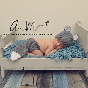 May include: A newborn baby wearing a gray and white crocheted wolf hat and matching pants sleeps on a miniature wooden bed with a blue fuzzy mattress. The bed is on a wooden floor.
