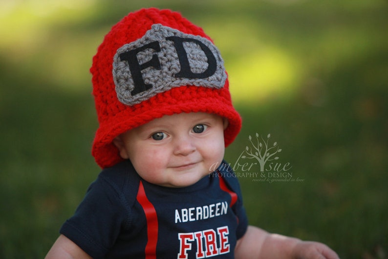 May include: A baby wearing a red crocheted firefighter hat with a gray "FD" patch and a navy blue shirt with the text "Aberdeen Fire".