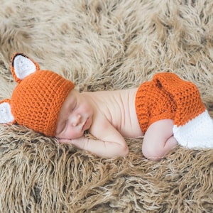 May include: A newborn baby wearing a crocheted fox costume, including a hat with ears and a diaper cover with a tail. The baby is sleeping on a brown fluffy blanket.
