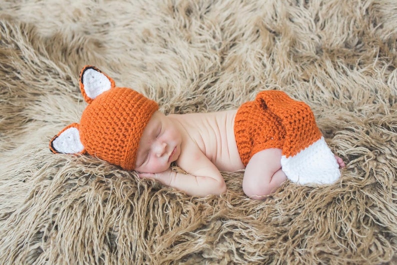 May include: A newborn baby wearing a crocheted fox costume, including a hat with ears and a diaper cover with a tail. The baby is sleeping on a brown fluffy blanket.