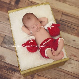 May include: A newborn baby wearing a red and white crocheted boxing outfit, lying on its back in a wicker basket lined with white faux fur. The baby is wearing a red and white crocheted boxing outfit with a red and white crocheted boxing glove on each hand. The baby is sleeping peacefully.