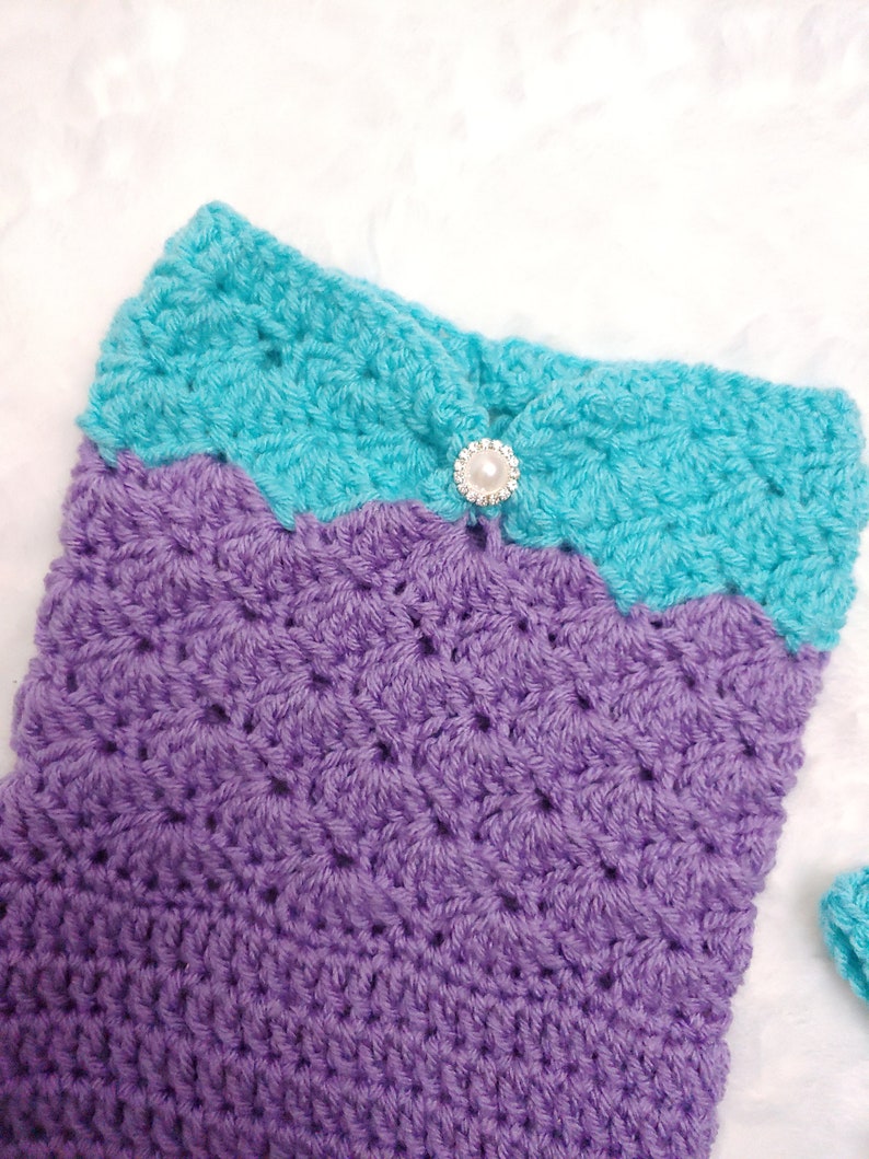 May include: A crocheted mermaid tail blanket in purple and turquoise with a pearl button.