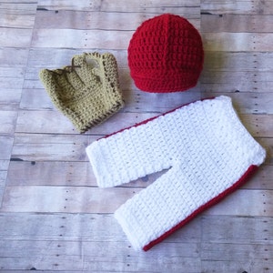 May include: A crocheted baby outfit in white and red with a matching red crocheted hat and a brown crocheted mitten.