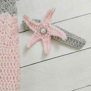 May include: A pink crocheted starfish with a pearl button in the center. The starfish is attached to a gray crocheted headband.