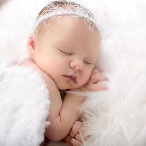 May include: A newborn baby sleeping peacefully on a white fluffy blanket, wearing a white crocheted headband.