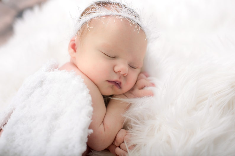 May include: A newborn baby sleeping peacefully on a white fluffy blanket, wearing a white crocheted headband.