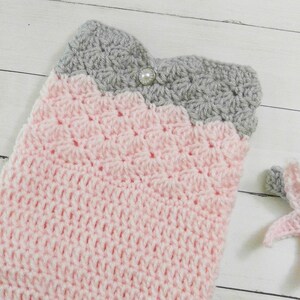 May include: A pink and gray crocheted baby blanket with a pearl button. The blanket is made with a textured stitch and has a scalloped edge.
