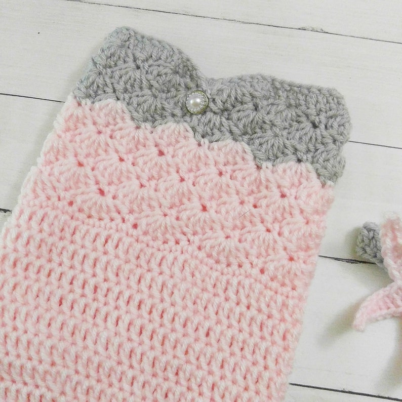 May include: A pink and gray crocheted baby blanket with a pearl button. The blanket is made with a textured stitch and has a scalloped edge.