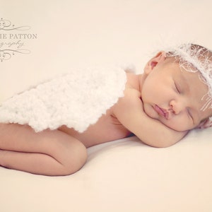 May include: A sleeping baby wearing a white crocheted headband and a white crocheted blanket. The baby is lying on its side with its head resting on its arm.
