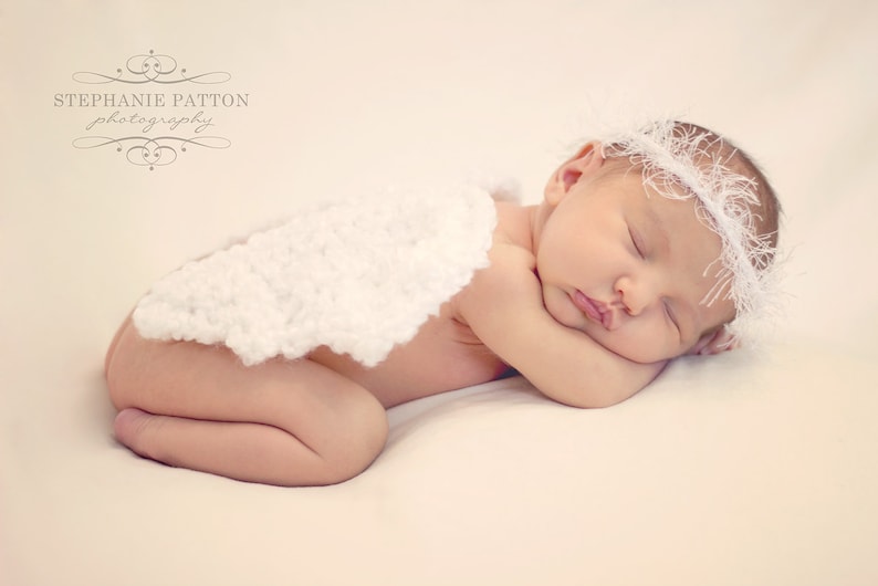 May include: A sleeping baby wearing a white crocheted headband and a white crocheted blanket. The baby is lying on its side with its head resting on its arm.