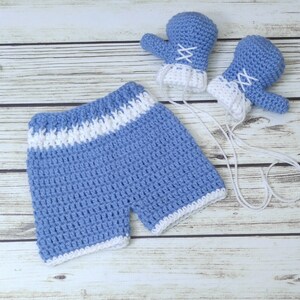 May include: A blue and white crocheted baby outfit with shorts and matching mittens. The outfit is made with a soft yarn and has a white trim.