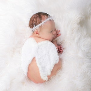 May include: A newborn baby wearing a white crocheted angel wing costume and a white crocheted headband, sleeping on a white fluffy blanket.