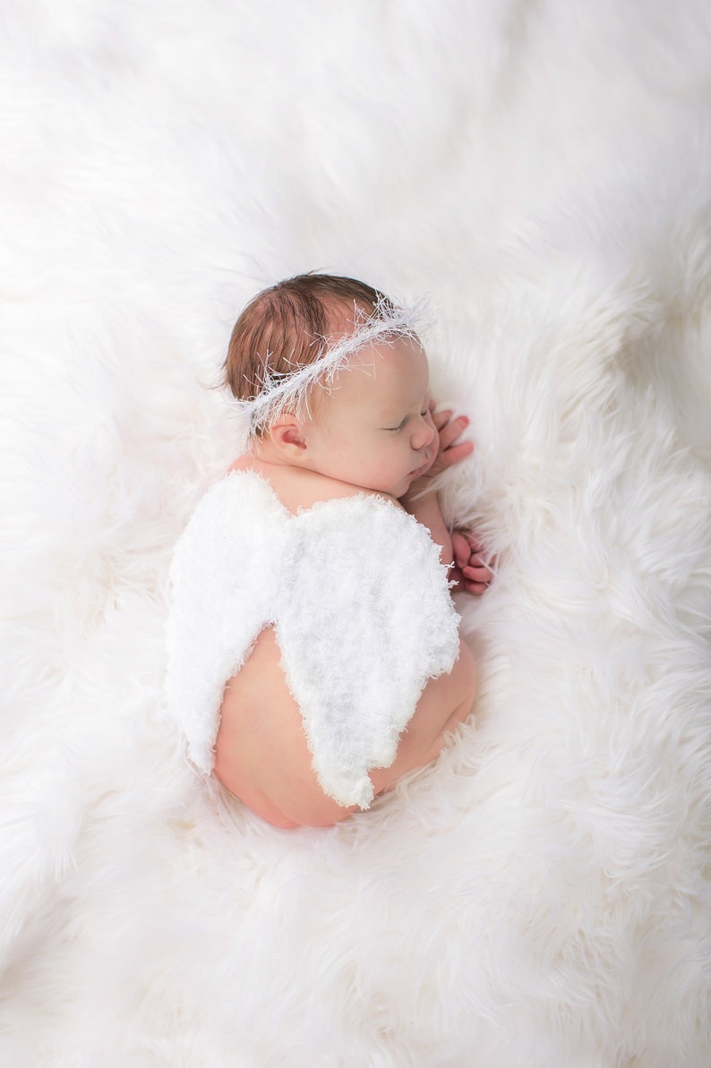 May include: A newborn baby wearing a white crocheted angel wing costume and a white crocheted headband, sleeping on a white fluffy blanket.