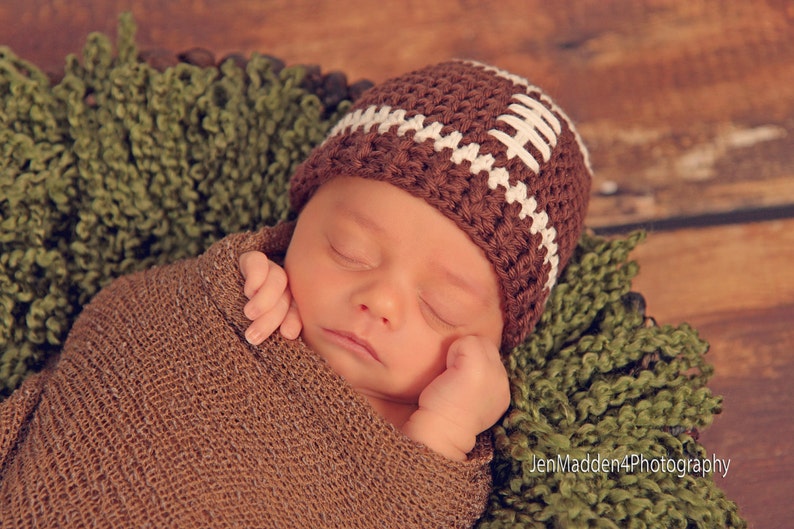 May include: A sleeping baby wearing a brown and white crocheted football hat. The baby is wrapped in a brown knitted blanket and is lying on a green fuzzy surface.